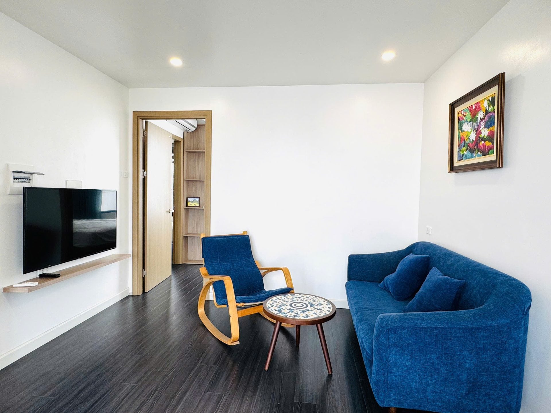 A living room with a blue sofa, a wooden rocking chair and a mosaic side table, and a colourful flower painting on the wall