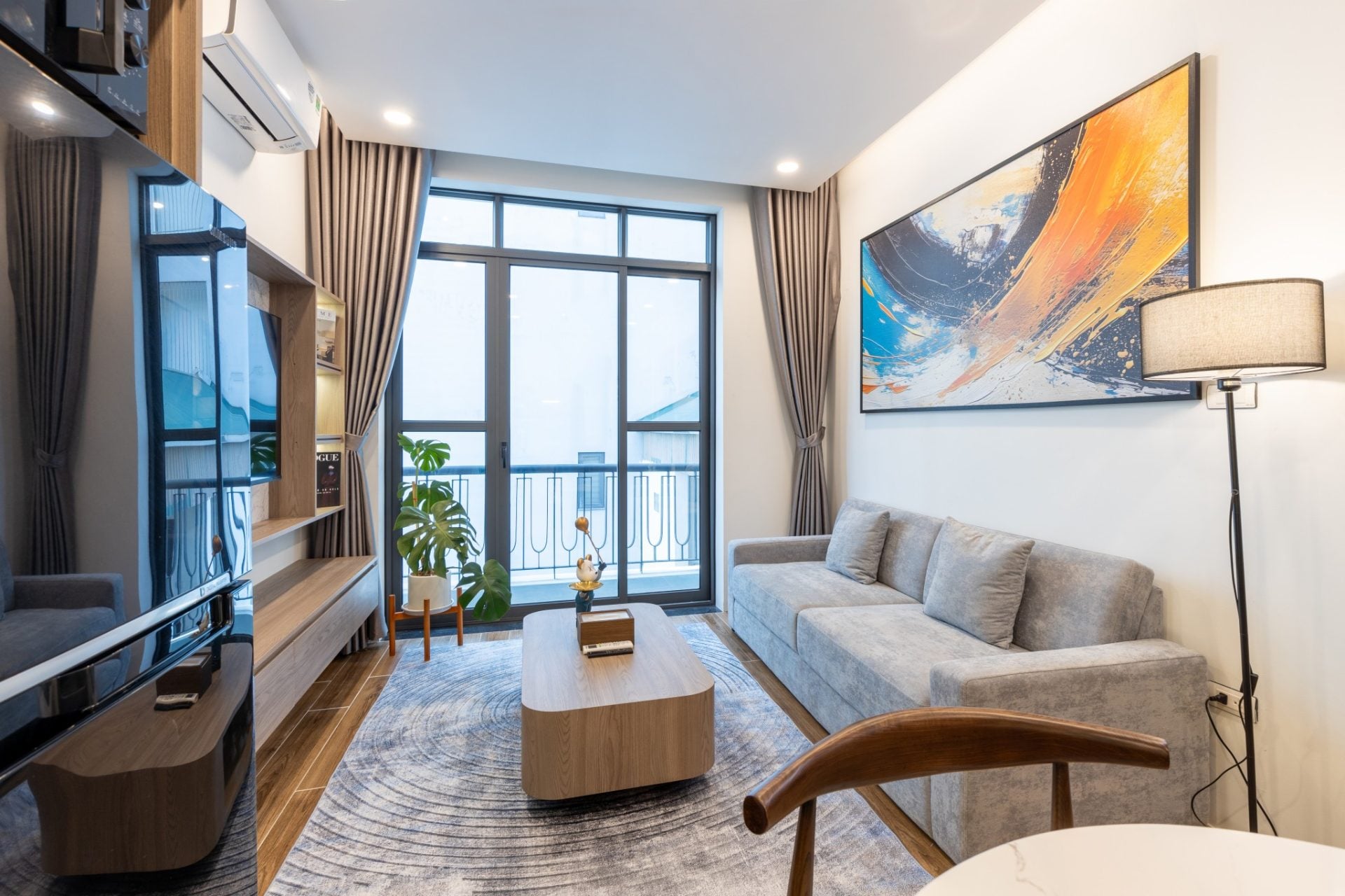 A furnished living room with a grey sofa, a wooden coffee table and a large abstract painting, with a balcony door beyond