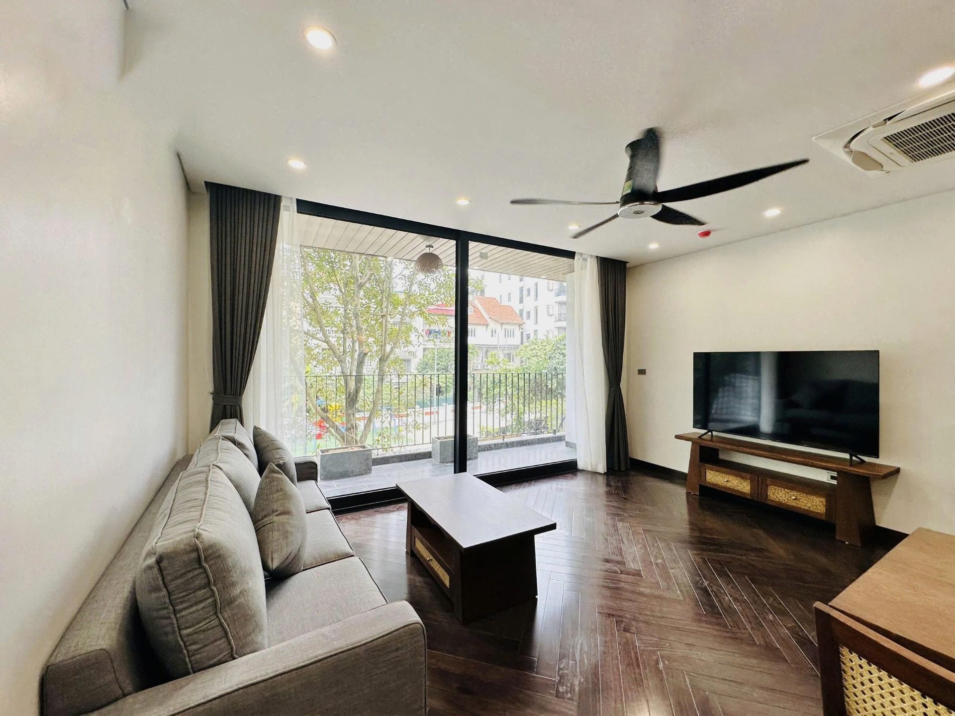 A living room with a grey sofa, a ceiling fan and dark herringbone floors, opening onto a balcony over trees