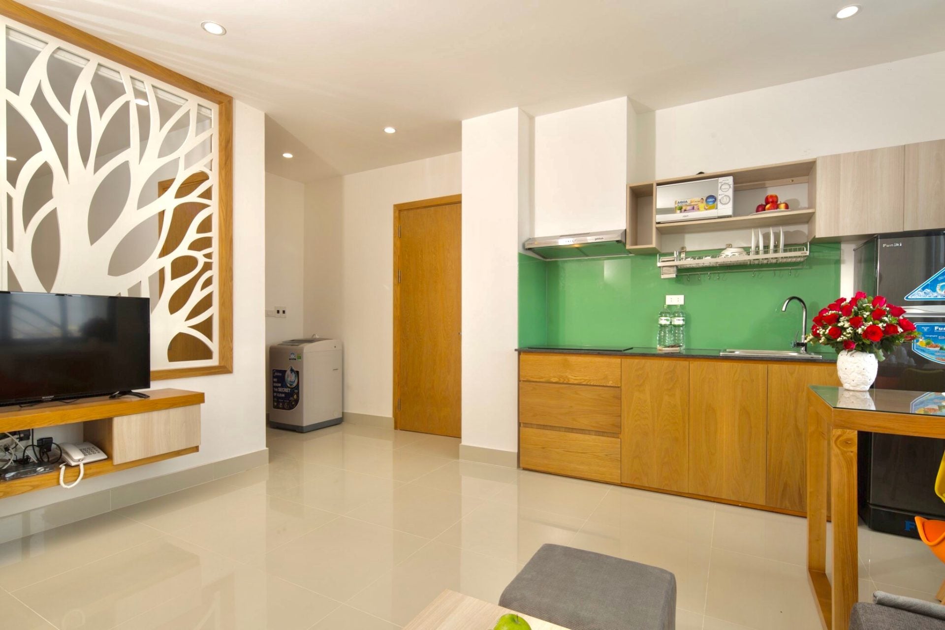 A bright one-bedroom apartment with a tree-pattern timber screen behind the television, a kitchen with a green glass splashback and a vase of red roses on the table
