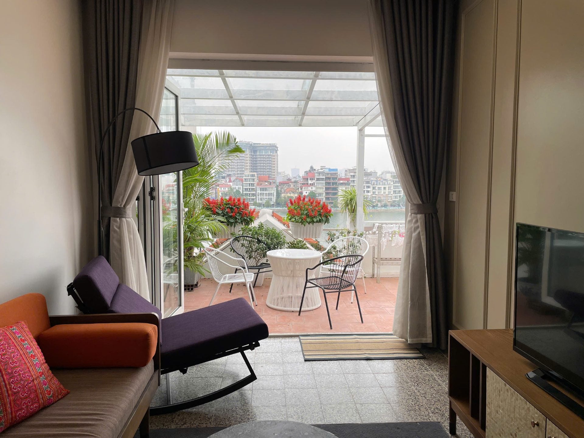 A living room opening onto a terrace with red flowers and white chairs, with West Lake and the city skyline beyond