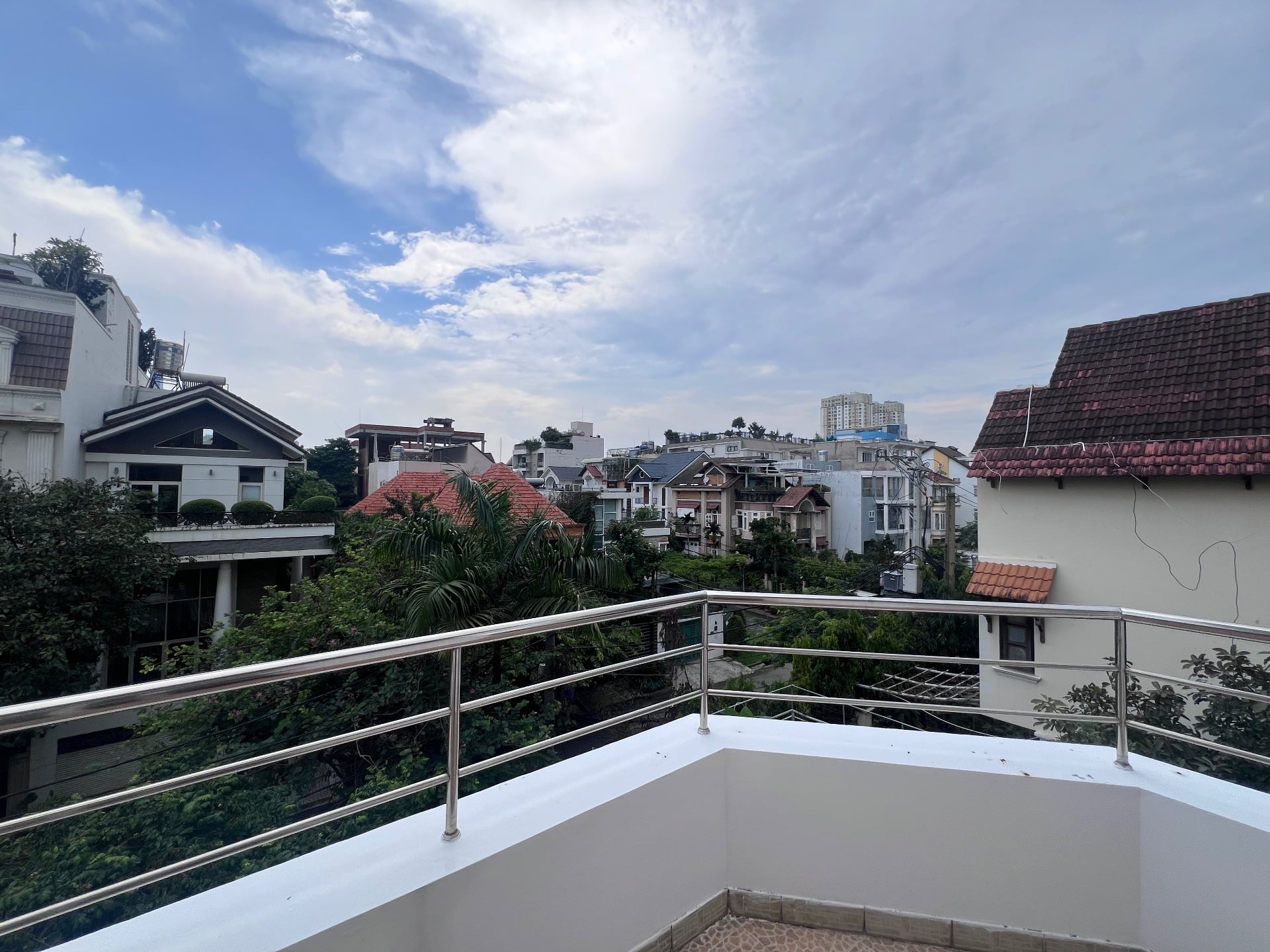 A balcony railing in Thảo Điền looking out over green treetops and the tiled rooftops of nearby villas under a blue sky