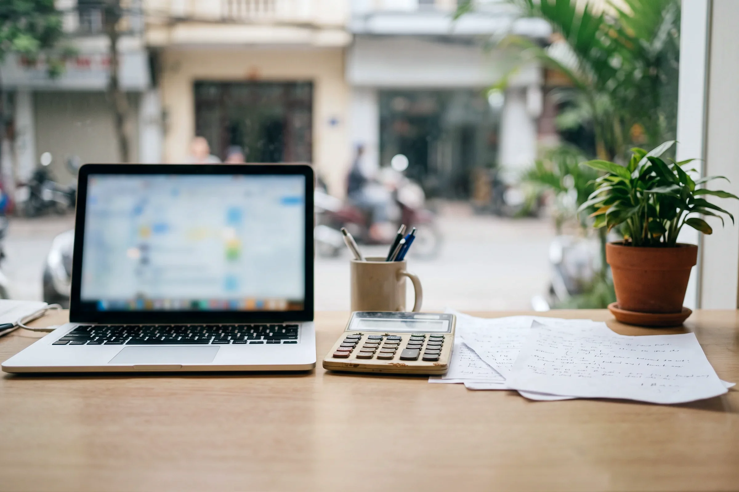 A laptop, a calculator and papers on a desk by a window
