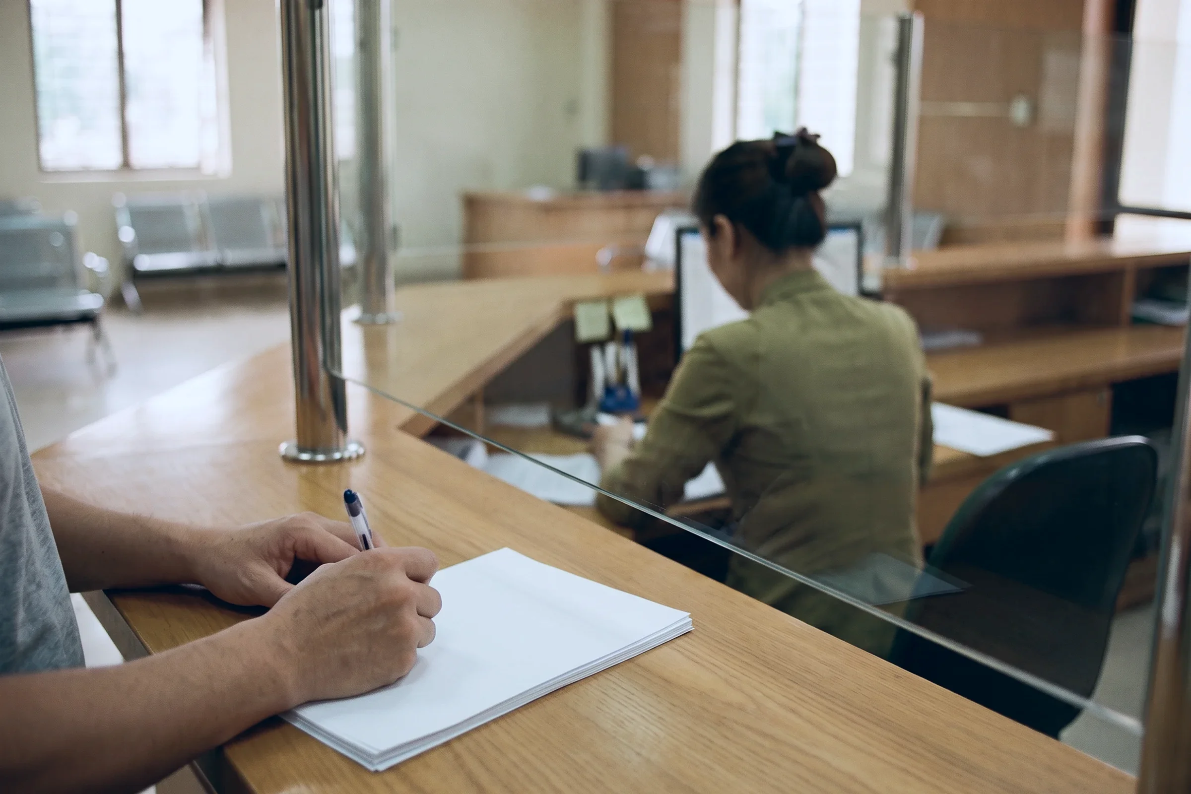 A hand filling in a form at a bank counter, the clerk out of focus behind