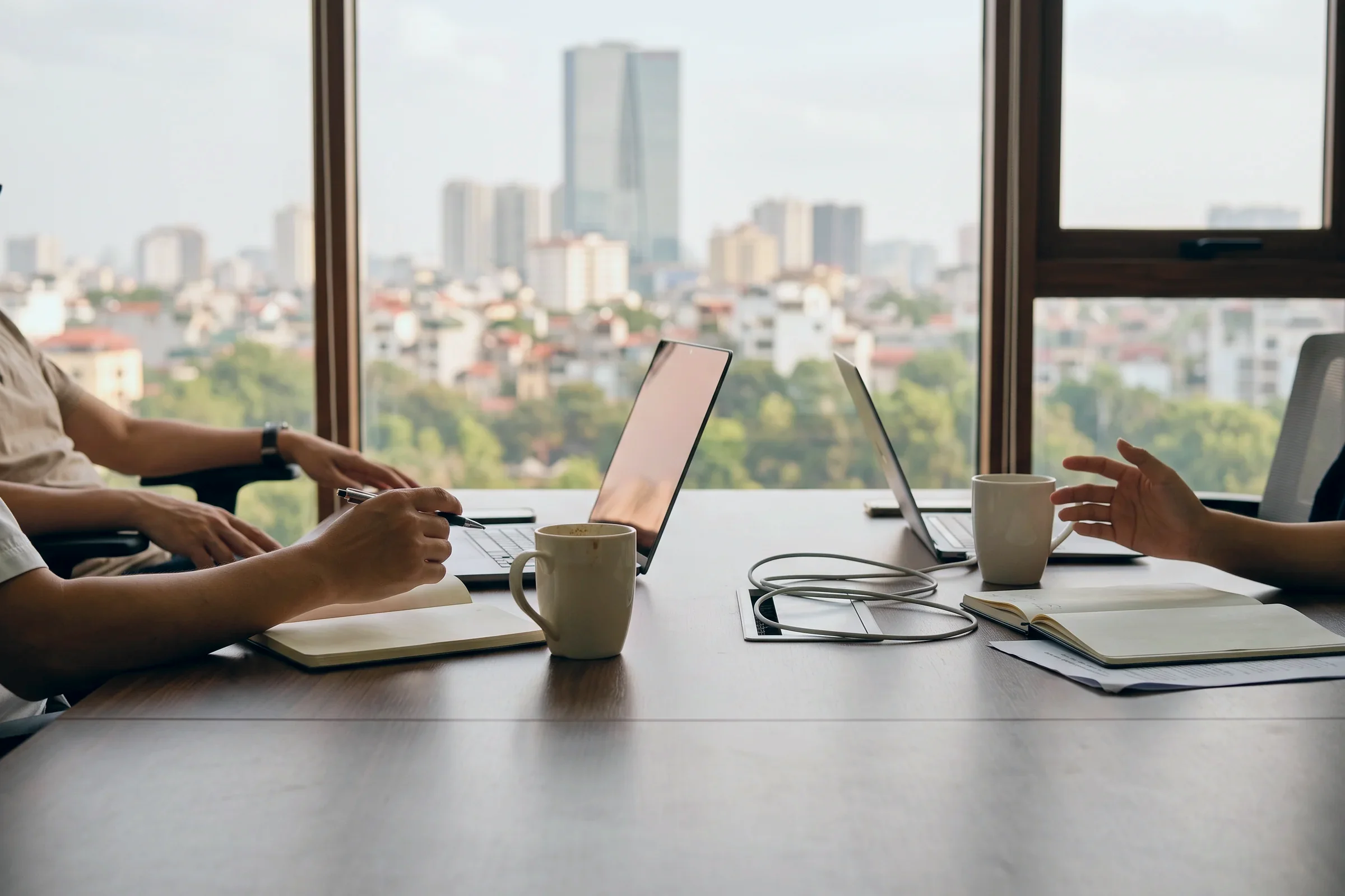 Two people at laptops at a meeting table, seen from the side, with the city through the window